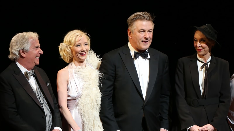 Four actors on stage wearing fancy outfits like tuxedos, and one woman wears a silver halter gown with a cream feather boa.
